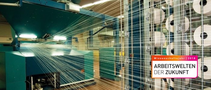 In einer Textilfabrik rollen 10 riesige Garnrollen ihr Garn maschinell und parallel ab in einen digital gesteuerten Webstuhl.