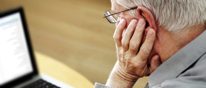 Ein älterer Herr mit grauem Haar und Brille stützt seinen Kopf auf seine linke Hand, während er auf einen Laptop schaut.