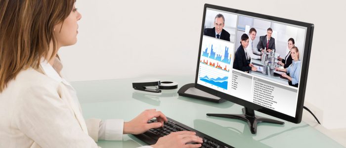 Eine Frau sitzt vor einem PC. Auf dem Bildschirm sind eine Konferenz und Grafiken zu sehen.