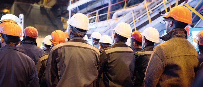 Eine Gruppe von Arbeitern mit Schutzhelmen werden von hinten vor einer Baustelle gezeigt