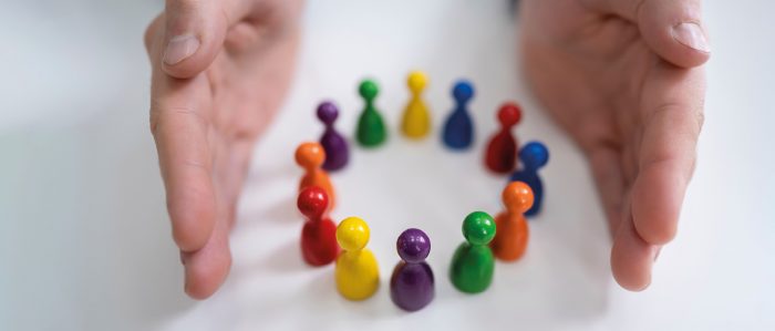 Spielfiguren in verschiedenen Farben sind in einem kleinen Kreis angeordnet. Zwei Handflächen befinden sich parallel zum Kreis und begrenzen ihn so nach außen.