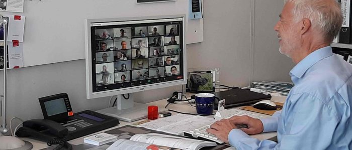 Lutz Bellmann sitzt in seinem Büro vor dem PC und nimmt an der Online-Tagung teil. Er tippt etwas mit der Tastatur und lächelt dabei. Auf dem Bildschirm sind die verschiedenen anderen Teilnehmerinnen und Teilnehmer der Online-Konferenz mit Kamerabild zu sehen.