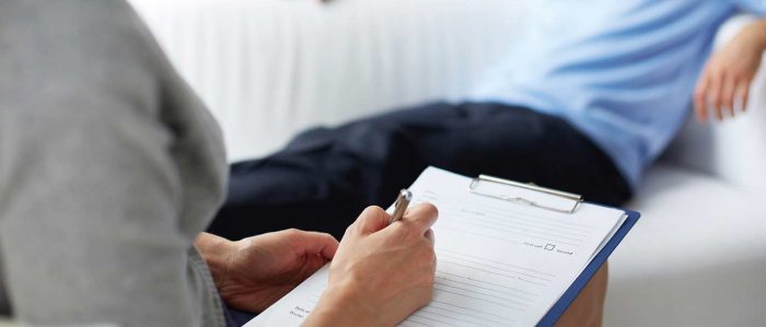 Mann sitzt auf einer Couch und blickt in die Ferne. Ihm gegenüber sitzt eine Therapeutin und schreibt auf ein Papier auf ihrem Klemmbrett.
