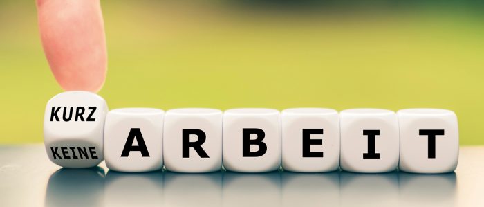 Würfelreihe, die das Wort „Arbeit“ ergibt. Vor dem Buchstaben „A“ befindet sich ein weiterer Würfel, der auf einer Seite das Wort „Kurz“, auf einer anderen Seite das Wort „Keine“ zeigt. Ein menschlicher Finger hält diesen Würfel in der Schwebe.