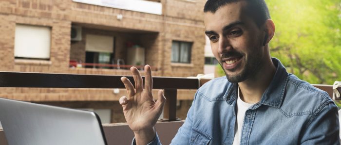 Ein Mann mit Migrationshintergrund sitzt vor einem Laptop und kommuniziert darüber virtuell. Er begrüßt sein Gegenüber, indem er die Hand hebt.