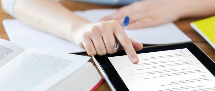 Frau sitzt mit Tablet und Buch am Schreibtisch.