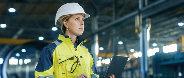 Junge Frau steht mit Arbeitsweste, Sicherheitshelm und Laptop in einer Werkshalle.