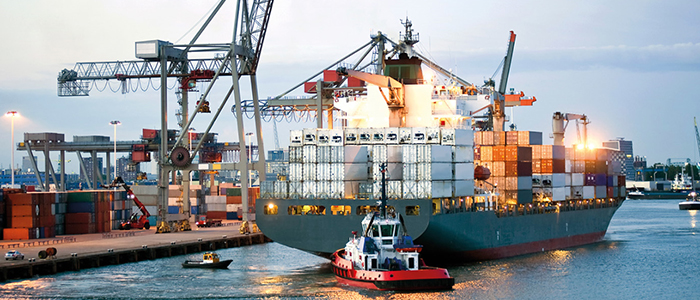 Das Bild zeigt ein mit Containern beladenes Schiff am Hafen. Es wird von zwei Begleitschiffen geführt.