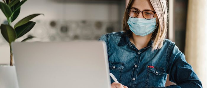 Frau mit Corona-Maske sitzt zu Haus am Laptop und schreibt etwas auf oder mit.