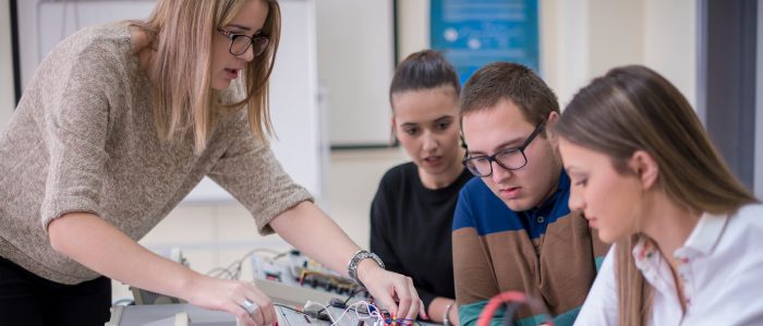 Drei Auszubildende bekommen von einer Mitarbeiterin eine technische Vorrichtung erklärt.