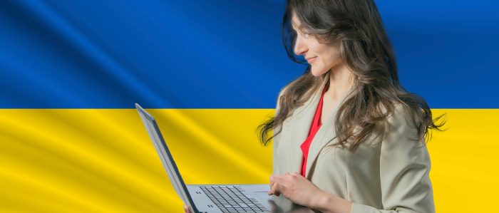 Eine junge Frau im Business-Look steht mit aufgeklapptem Laptop vor einer ukrainischen Flagge.
