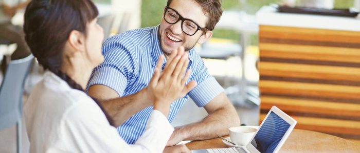 Eine Frau und ein Mann sitzen zusammen an einem Tisch vor einem Laptop und schlagen lachend mit den Händen ein.
