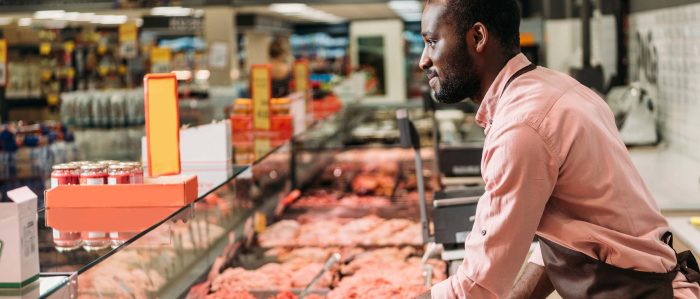 Ein Mann mit schwarzer Hautfarbe steht hinter einer Fleischtheke im Supermarkt als Verkäufer.