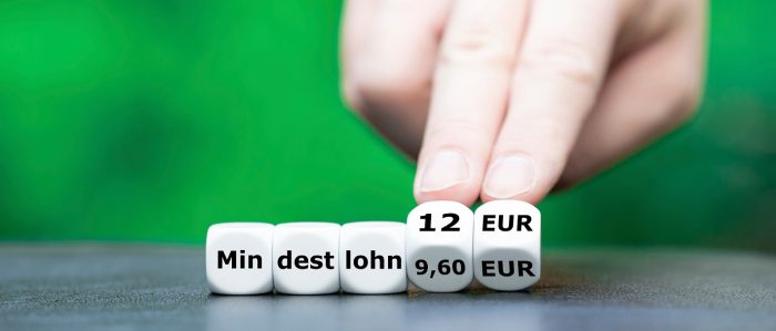 5 weiße Würfel bilden zusammen das Wort Mindestlohn. Eine menschliche Hand dreht die beiden letzten Würfel von 9,60 auf 12 Euro.