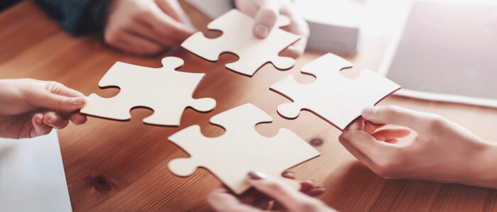 Die Aufnahme von schräg oben zeigt vier Hände von Personen in Geschäftskleidung, die gerade vier Puzzle-Teile über einem Holztisch zusammenfügen.