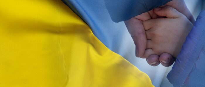 Das Bild zeigt eine Mutter, die die Hand ihres Kindes hält vor dem Hintergrund einer ukrainischen Flagge.