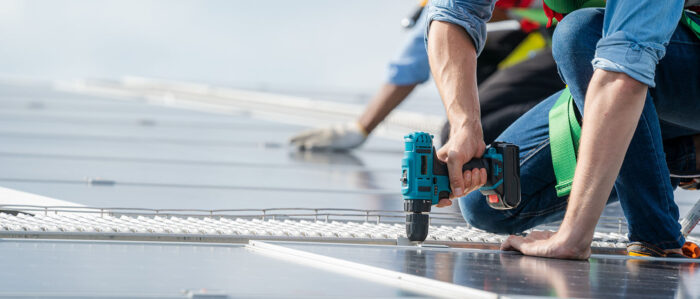Ein Handwerker installiert mit einer Bohrmaschine Solarpanele auf einem Gebäudedach.