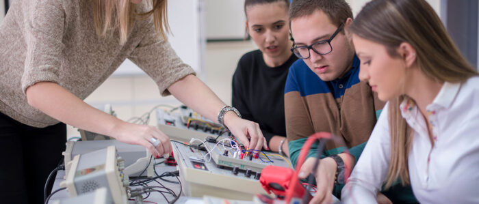Eine Frau nutzt elektrotechnische Geräte und zwei junge Frauen und ein junger Mann sehen dabei zu