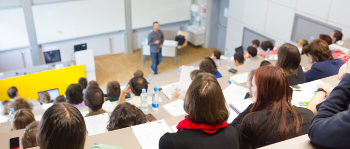 Blick von hinten oben in einen mit Studierenden gefüllten Hörsal, in dem gerade eine Vorlesung stattfindet.