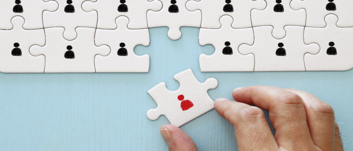 Eine Hand fügt ein Puzzlestück mit einer roten Figur in ein Puzzle mit vielen schwarzen Figuren ein.