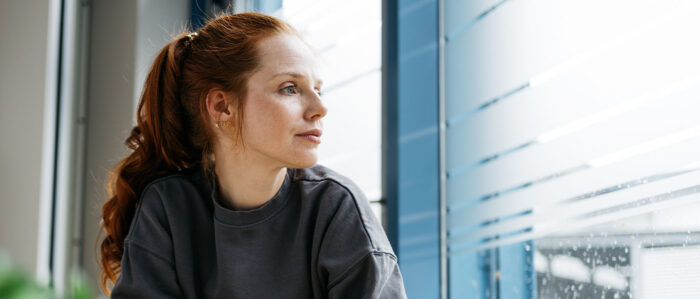 A young woman sits at a table by the window and looks out, lost in thought. On the table are a cup, a cell phone and a potted plant.