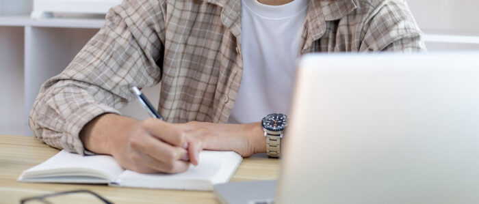Mann sitzt am Schreibtisch und macht sich Notizen in einen Schreibblock, während er auf den Laptop vor ihm schaut. Nahaufnahme auf den Oberkörper und Arme.
