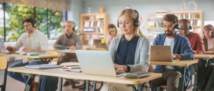 Adult men and women of different ages each sit at a desk in a training room and work with headsets on their laptops.