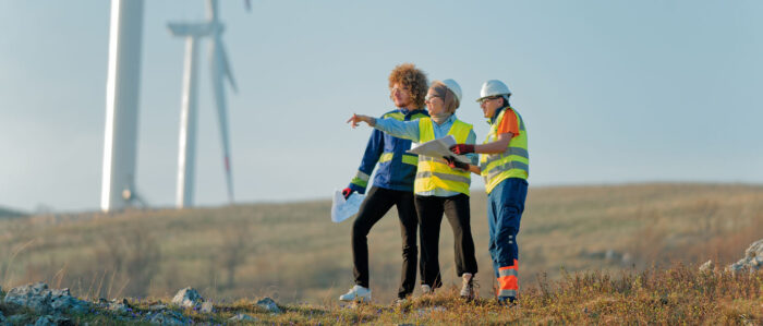 Two men and a woman, all wearing high-visibility waistcoats, are standing in a modern wind farm and planing a project.