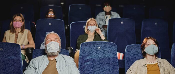 Frontalaufnahme von mehreren Männern und Frauen, die sich einen Film im Kino ansehen. Sie tragen alle einen Mundschutz und zwischen ihnen ist jeweils ein Sitz frei.