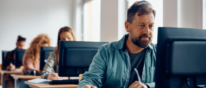 Perspektivische Aufnahme von erwachsenen Kursteilnehmern, die in einer Reihe hintereinander an ihren mit Computern ausgestatteten Schreibtischen arbeiten. Im Fokus ist der Mann, der vorne sitzt.