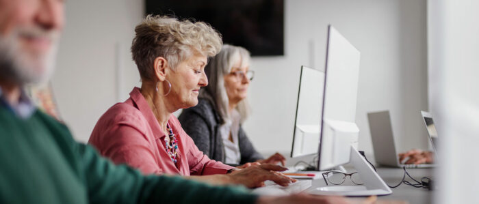 Perspektivische Aufnahme von erwachsenen Kursteilnehmern, die nebeneinander in einer Reihe an Computern arbeiten. Im Fokus ist eine Frau, die in der Mitte sitzt.
