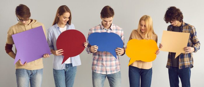 Eine Gruppe Jugendlicher steht vor einem grauem Studiohintergrund. Die Mädchen und Jungen schauen auf die bunten, leeren Sprechblasen aus Pappe, die sie in den Händen halten.