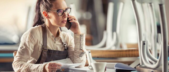 Junge Besitzerin einer Gastronomie sitzt in Ihrem geschlossenen Restaurant bei einem Kaffee über Ihren Unterlagen während sie mit dem Handy telefoniert.