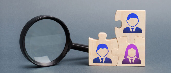 Three interlocking puzzle pieces, each with a symbol for a person, are in front of a magnifying glass.