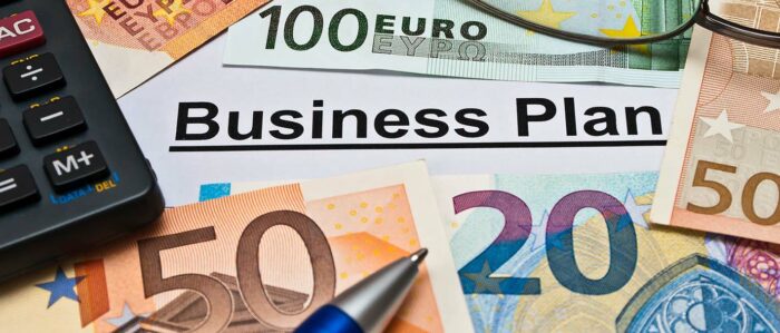 Nahaufnahme von einem Business-Plan, der größtenteils von Euro-Scheinen, einem Kugelschreiber, einer Brille und einem Taschenrechner verdeckt wird.