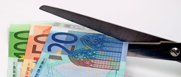 Nahaufnahme einer Schere, wie sie einen 20-Euro-Schein, einen 50-Euro-Schein und einen 100-Euro-Schein durchschneidet.