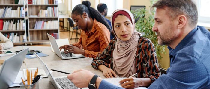 A teacher explains something to an female adult Muslim student wearing a hijab on the laptop they are both sitting in front of.