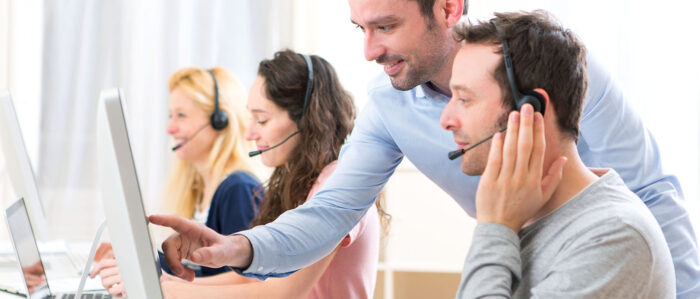 Training situation for call center employees: two women and a man are sitting in front of their computers with headsets. A man in a shirt stands behind the man with the headset and points at his monitor. Everyone looks friendly.