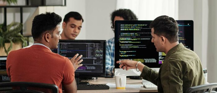 Vier junge Softwareentwickler sitzen an ihren Rechnern und diskutieren. Auf den Monitoren sind Programmiercodes erkennbar.
